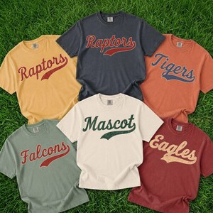 A collection of six t-shirts with different team names printed on them, laid out on a grassy surface.