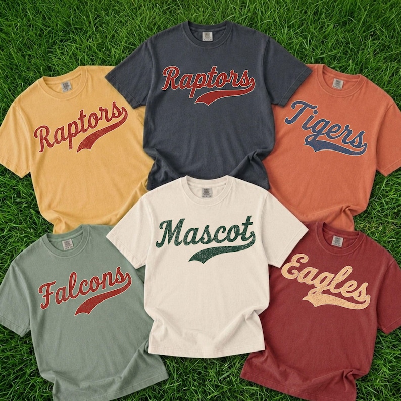 A collection of six t-shirts with different team names printed on them, laid out on a grassy surface.