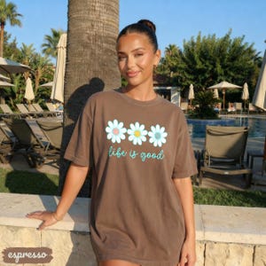 Puede incluir: Camiseta color espresso con tres gr&aacute;ficos de margaritas con centros rosas, amarillos y azules. Las palabras "life is good" est&aacute;n escritas en una fuente azul claro debajo de las flores. La camiseta la lleva una persona.