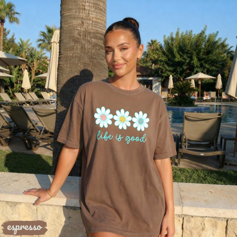 Puede incluir: Camiseta color espresso con tres gr&aacute;ficos de margaritas con centros rosas, amarillos y azules. Las palabras "life is good" est&aacute;n escritas en una fuente azul claro debajo de las flores. La camiseta la lleva una persona.