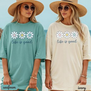 Puede incluir: Dos camisetas holgadas, una verde mar y otra marfil, cada una con un dise&ntilde;o de margarita y el texto "life is good". Las camisetas tienen mangas cortas y se usan con sombreros de paja y gafas de sol. La camiseta verde mar est&aacute; a la izquierda y la marfil a la derecha.