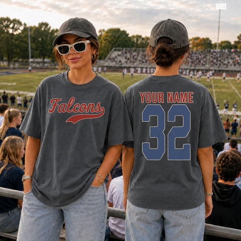 A woman wearing a grey t-shirt with the word "FALCONS" and the number "32" on it, standing in a stadium with a crowd of people in the background.