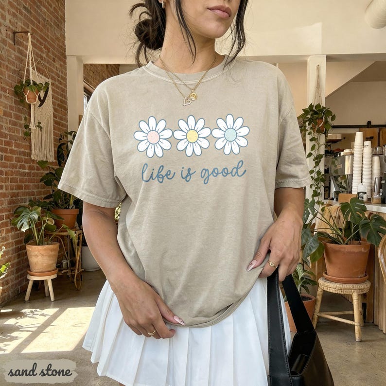 Puede incluir: Camiseta color arena con un gr&aacute;fico de margaritas y la frase "Life is good" en una fuente cursiva. El gr&aacute;fico de la margarita tiene centros rosas, amarillos y azules. La camiseta es de un estilo casual y c&oacute;modo.