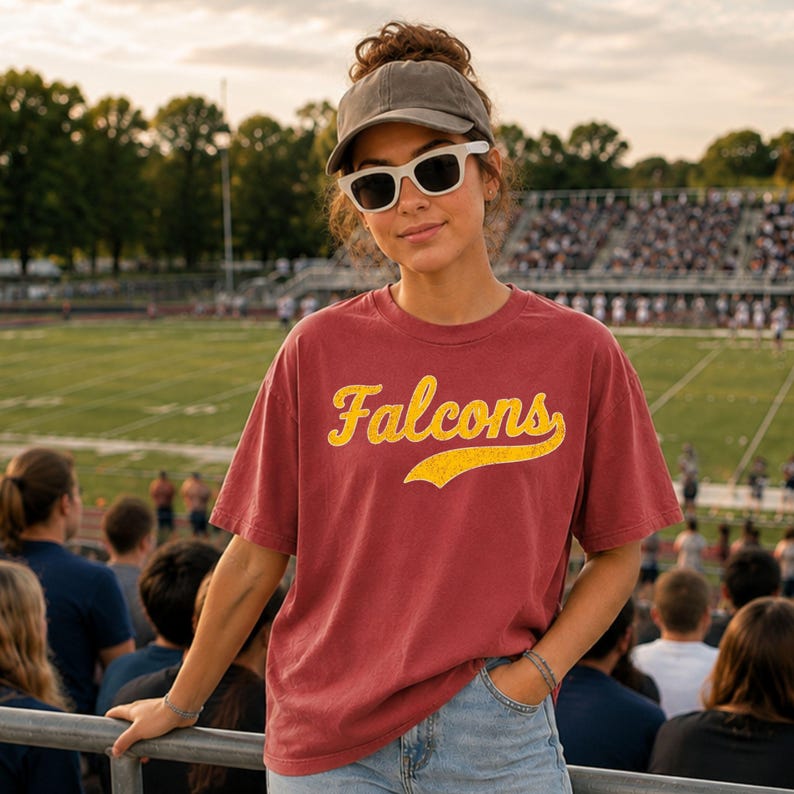 A young woman wearing a red t-shirt with the word "FALCONS" stands in a stadium, smiling and leaning on a railing.