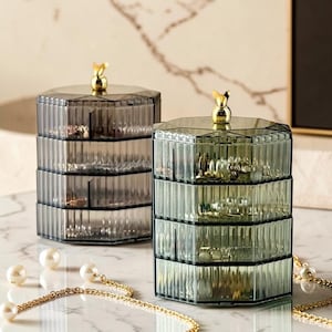 May include: Two hexagonal, multi-tiered storage containers made of translucent ribbed plastic. One container is dark gray and the other is olive green. Each features four rotating compartments and a small, gold-colored animal-shaped handle on top.