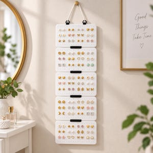 May include: A white wall-mounted earring organizer featuring five rectangular panels connected by black clips. The panels are perforated with numerous small holes, each holding a pair of stud earrings in various shapes like stars, hearts, and flowers.