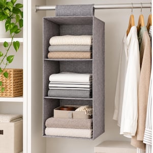 May include: A gray fabric hanging closet organizer with three open shelves suspended from a metal rod. The shelves contain folded sweaters, shirts, and a small beige storage bin. The organizer features a textured, heathered gray material.