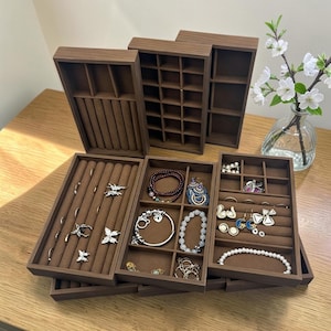 May include: A set of brown wooden jewelry organizer trays with various compartments, including ring rolls and grid sections. The trays are filled with assorted jewelry pieces such as bracelets, earrings, and rings, showcasing a functional storage solution for organizing accessories.