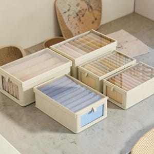 May include: Four rectangular beige fabric storage organizers with clear plastic lids and front windows. Each box contains multiple internal dividers holding neatly folded textiles in shades of blue, beige, and tan, arranged in rows.