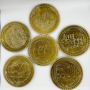 May include: A set of five circular olive-green ceramic plates, each featuring a raised relief design of a different French castle. The plates include embossed labels for Saumur, Villandry, Loches, and other locations, with scalloped edges.