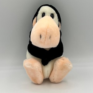 Pode incluir: Um brinquedo de pelúcia de pinguim com cabeça e cachecol pretos, corpo creme e pés e bico cor de pêssego. O pinguim está sentado em posição vertical. O brinquedo tem olhos pretos de botão.