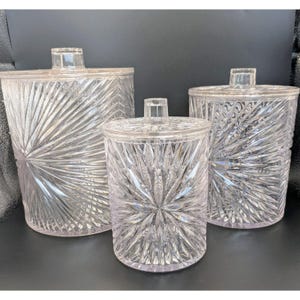 May include: A set of three clear acrylic canisters with lids, each featuring a textured sunburst pattern radiating from the center. The cylindrical containers vary in size, with one large canister, one medium canister, and one small canister.