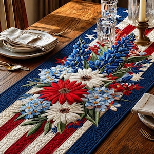 May include: A patriotic table runner with a floral design. The runner features red and white stripes, blue borders with white stars, and embroidered flowers in red, white, and blue. The runner is placed on a wooden table.