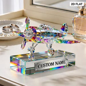 May include: A decorative crystal airplane with a rainbow prism effect. The airplane sits on a rectangular base with a silver plate that reads "CUSTOM NAME". The propeller, wings, and body of the plane are made of faceted crystal.