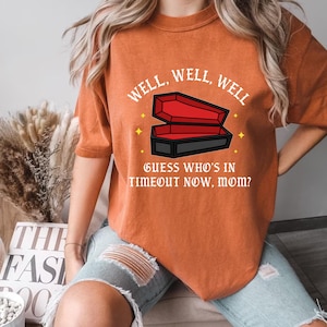 May include: Orange t-shirt with the text "WELL, WELL, WELL" above a coffin graphic. Below the coffin, the text reads "GUESS WHO'S IN TIMEOUT NOW, MOM?" The shirt is a casual style.