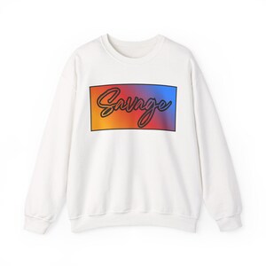 Puede incluir: Sudadera blanca de cuello redondo con la palabra "Savage" en cursiva, dentro de un rectángulo degradado naranja, rojo y azul. Una sudadera de estilo casual.
