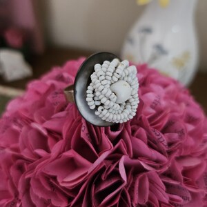 May include: A handcrafted ring featuring a dark gray, circular setting adorned with a cluster of small, white beads in a floral design. The ring is set on a pink, paper flower arrangement, creating a unique and artistic aesthetic.