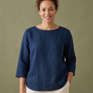 Puede incluir: Una blusa de lino azul marino con mangas tres cuartos. La blusa tiene un cuello redondo y un corte holgado. La modelo lleva la blusa con pantalones color crema. El fondo es verde oliva.