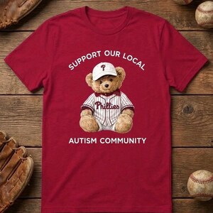 May include: A red t-shirt with the text "SUPPORT OUR LOCAL AUTISM COMMUNITY." The shirt features a teddy bear wearing a baseball uniform and a white cap with a "P" logo. Baseball-themed items are in the background.