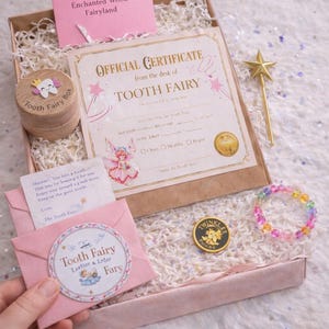 May include: A Tooth Fairy gift set in a box includes a certificate, a wooden tooth box, a gold fairy coin, a beaded bracelet, and a pink envelope with a Tooth Fairy sticker. The set has a magical theme with pink and gold accents.