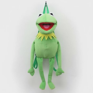 May include: A green plush backpack shaped like a frog with a wide open mouth, red tongue, and yellow collar. The backpack has green straps and is hanging against a white background.