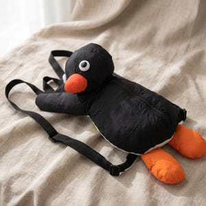 Könnte beinhalten: Ein schwarzer, pinguinförmiger Rucksack mit orangefarbenen Füßen und Schnabel sowie weißen Augen. Der Rucksack hat verstellbare schwarze Träger und einen Reißverschluss. Der Pinguin liegt auf einer beigen Oberfläche.