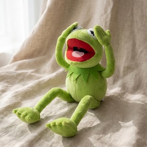 May include: A plush Kermit the Frog toy in a seated position with arms raised. The frog is light green with a red mouth, white eyes, and a green collar. The toy is on a beige fabric surface.