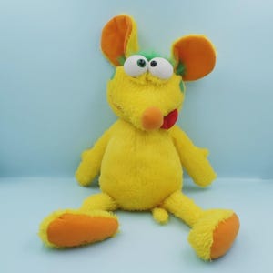 Puede incluir: Un peluche amarillo con forma de ratón con grandes orejas naranjas, una nariz redonda naranja y una lengua roja que sobresale. El juguete tiene manchas verdes y ojos grandes y blancos. El ratón está sentado con las piernas separadas y los pies naranjas.