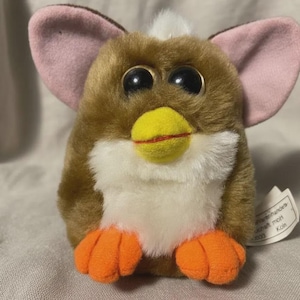 Puede incluir: Un juguete de peluche Furby marrón, blanco, amarillo, rosa y naranja. El Furby tiene ojos negros grandes, un pico amarillo con una línea roja y pies naranjas. Las orejas son marrones con el interior rosa. Juguete retro coleccionable.