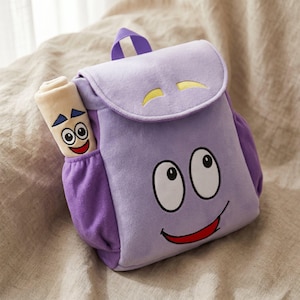 May include: A plush purple backpack featuring a large cartoon face with white eyes and a red smile on the front flap and main body. A rolled-up beige map character with blue eyebrows and a smiling face is tucked into the side pocket of the bag.