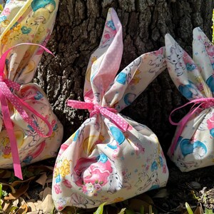 May include: Three Easter-themed fabric gift bags with bunny ear tops. The bags are tied with pink ribbons and feature a colorful print of bunnies, chicks, and flowers. The background includes a tree trunk and foliage.