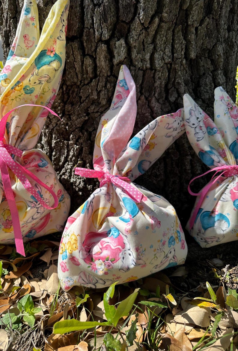 May include: Three Easter-themed fabric gift bags with bunny ear tops. The bags are tied with pink ribbons and feature a colorful print of bunnies, chicks, and flowers. The background includes a tree trunk and foliage.