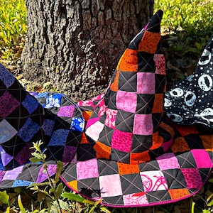 K&ouml;nnte beinhalten: Drei handgefertigte Hexenh&uuml;te mit gesteppten Patchwork-Designs. Ein Hut hat einen schwarzen Hintergrund mit rosa, orangefarbenen und schwarzen Quadraten. Ein anderer Hut hat einen schwarzen Hintergrund mit blauen, lila und schwarzen Quadraten. Der dritte Hut ist schwarz mit wei&szlig;en Monden und Sternen.