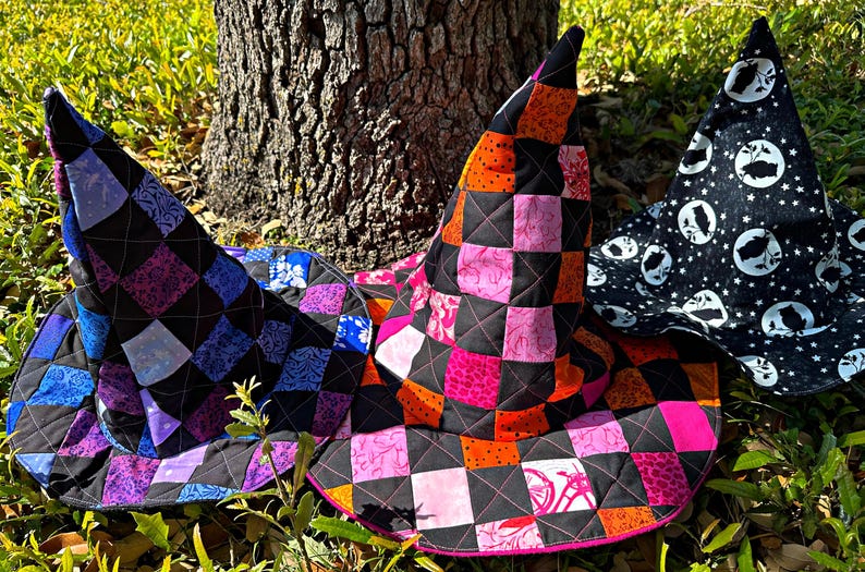 K&ouml;nnte beinhalten: Drei handgefertigte Hexenh&uuml;te mit gesteppten Patchwork-Designs. Ein Hut hat einen schwarzen Hintergrund mit rosa, orangefarbenen und schwarzen Quadraten. Ein anderer Hut hat einen schwarzen Hintergrund mit blauen, lila und schwarzen Quadraten. Der dritte Hut ist schwarz mit wei&szlig;en Monden und Sternen.