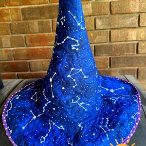 May include: A quilted witch hat in deep blue fabric featuring a white celestial constellation pattern. The hat has a tall, pointed crown and a wide brim finished with a purple and white polka dot fabric trim along the outer edge.
