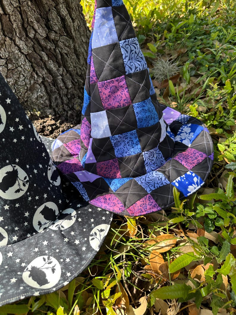 K&ouml;nnte beinhalten: Ein gesteppter Hexenhut mit einem Patchwork-Design in Lila, Blau und Schwarz. Der Hut besteht aus Stoffquadraten, die mit wei&szlig;en N&auml;hten zusammengen&auml;ht sind. Ein zweiter Hut mit einem schwarz-wei&szlig;en Mond- und Sternenmuster ist ebenfalls zu sehen.