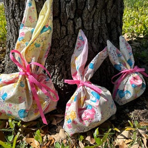 May include: Three Easter-themed gift bags shaped like bunnies, tied with pink ribbons. The bags are made of fabric with a pastel pattern of bunnies, flowers, and other Easter-related images. The bunny ears are pink.