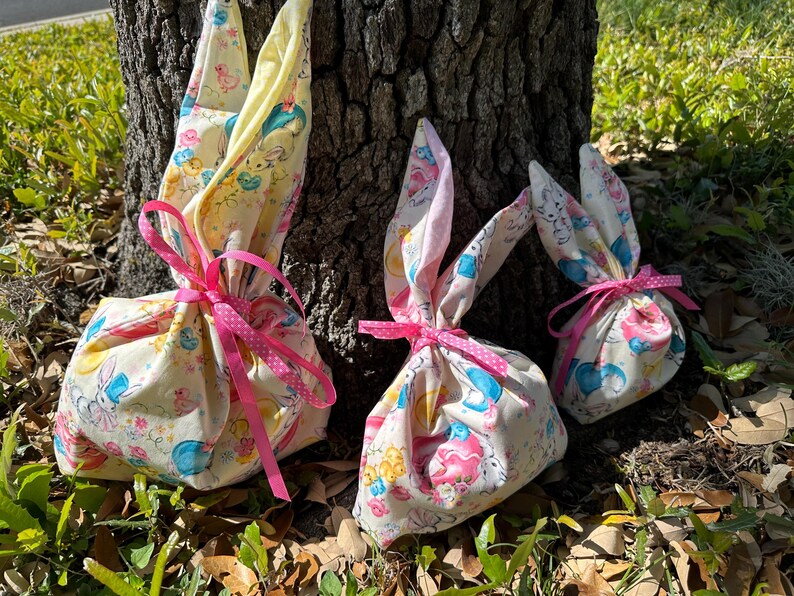 May include: Three Easter-themed gift bags shaped like bunnies, tied with pink ribbons. The bags are made of fabric with a pastel pattern of bunnies, flowers, and other Easter-related images. The bunny ears are pink.