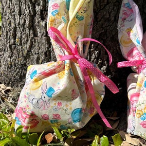 May include: Two Easter-themed gift bags with bunny ears. The bags are made of a light yellow fabric with a pastel bunny and chick pattern. They are tied with pink ribbons and sit on a bed of green leaves.