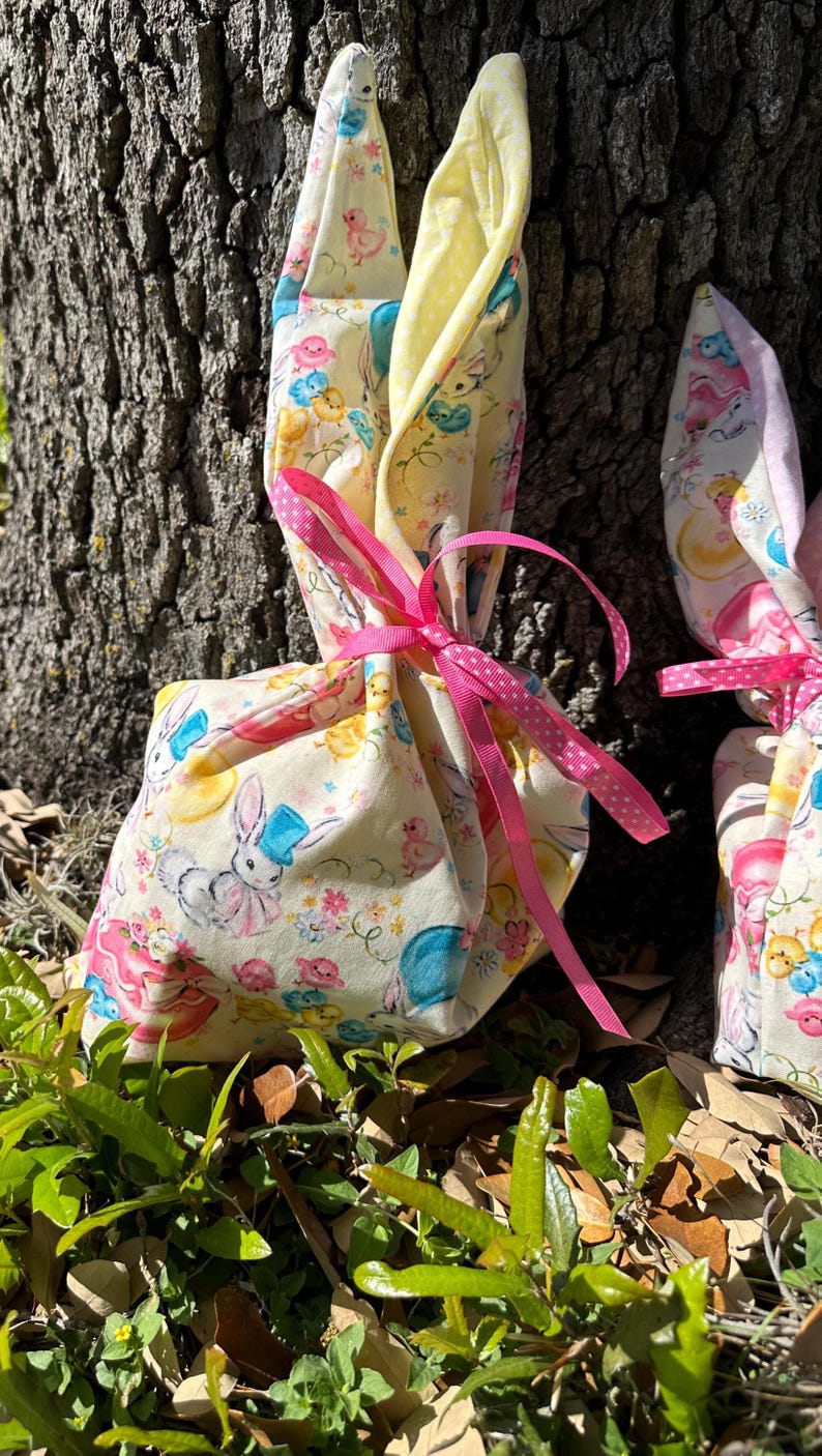 May include: Two Easter-themed gift bags with bunny ears. The bags are made of a light yellow fabric with a pastel bunny and chick pattern. They are tied with pink ribbons and sit on a bed of green leaves.