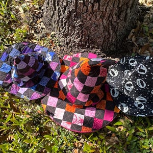 K&ouml;nnte beinhalten: Drei bunte, gesteppte Hexenh&uuml;te liegen auf dem Boden in der N&auml;he eines Baumstamms. Die H&uuml;te weisen Patchwork-Designs in verschiedenen Farben auf, darunter Lila, Rosa, Orange und Schwarz. Ein Hut ist schwarz mit wei&szlig;en Geistersilhouetten.