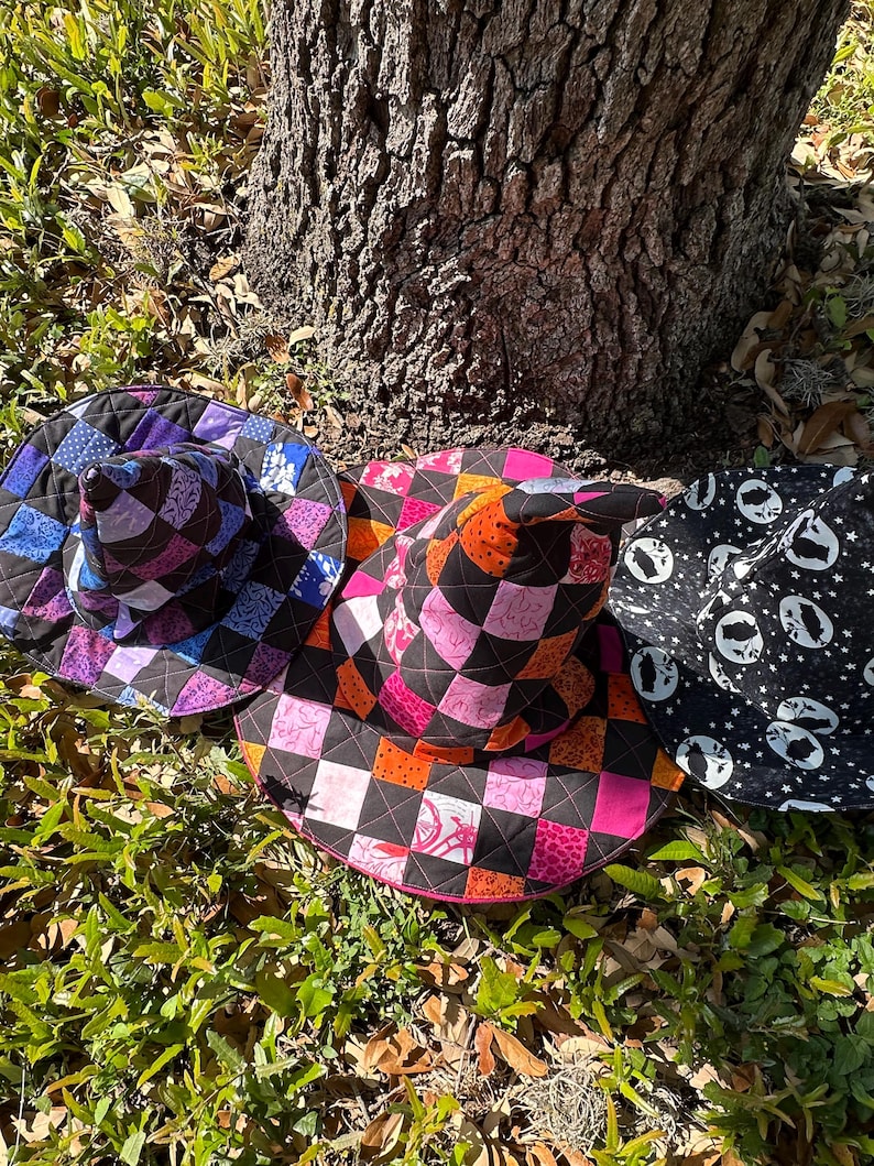 K&ouml;nnte beinhalten: Drei bunte, gesteppte Hexenh&uuml;te liegen auf dem Boden in der N&auml;he eines Baumstamms. Die H&uuml;te weisen Patchwork-Designs in verschiedenen Farben auf, darunter Lila, Rosa, Orange und Schwarz. Ein Hut ist schwarz mit wei&szlig;en Geistersilhouetten.