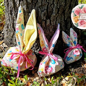 May include: Three Easter-themed fabric gift bags shaped like bunnies, tied with pink polka-dot ribbons. The bags are decorated with a pastel pattern of bunnies, ducks, and other spring motifs. The bunny ears are yellow and pink.