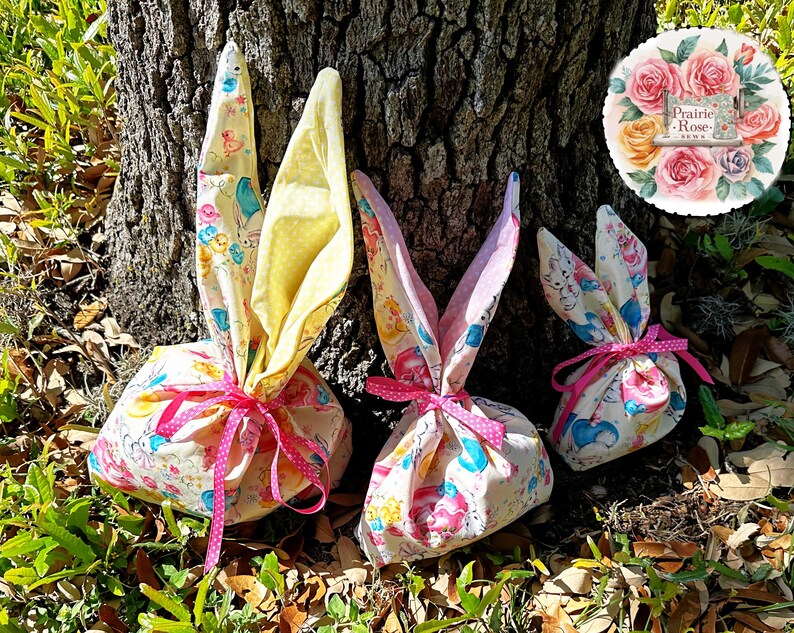 May include: Three Easter-themed fabric gift bags shaped like bunnies, tied with pink polka-dot ribbons. The bags are decorated with a pastel pattern of bunnies, ducks, and other spring motifs. The bunny ears are yellow and pink.