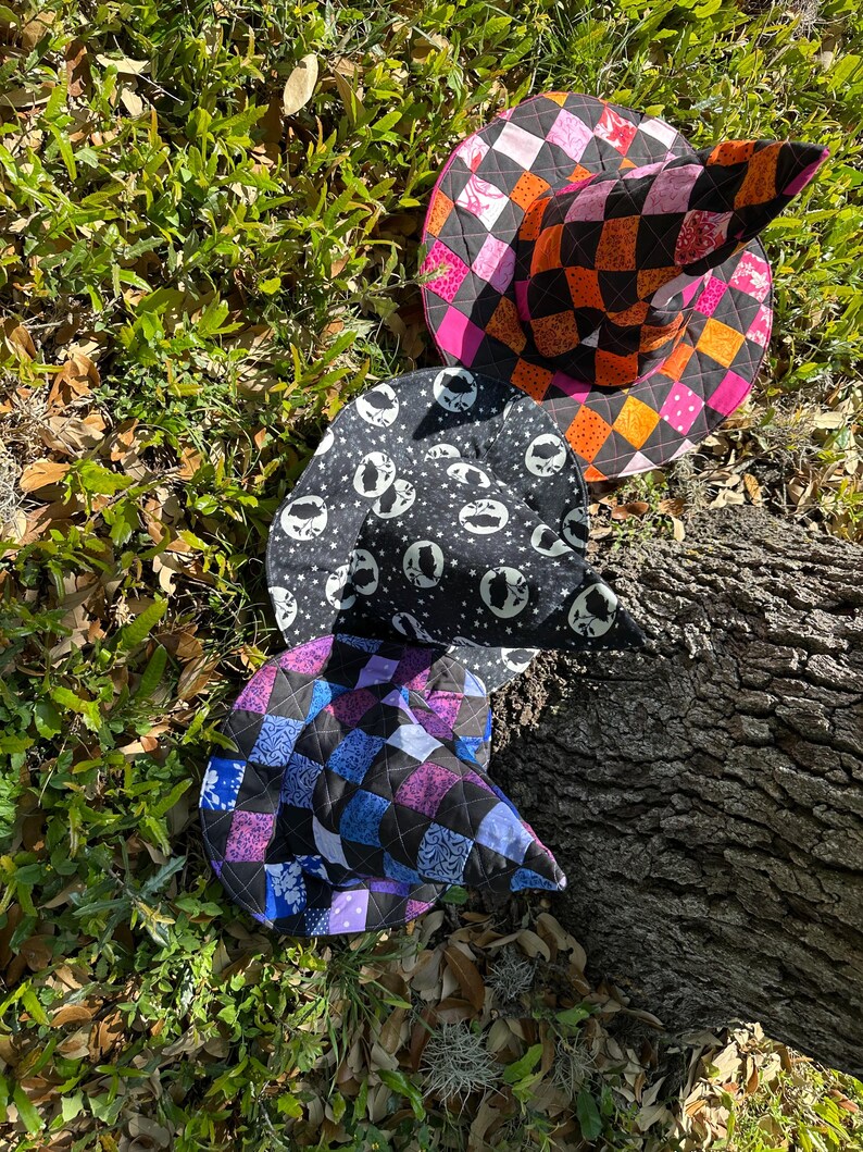 K&ouml;nnte beinhalten: Drei gesteppte Hexenh&uuml;te auf Gras und einem Baumstamm. Ein Hut hat ein schwarz-oranges Schachbrettmuster, ein anderer einen schwarz-wei&szlig;en Totenkopf-Druck, und der dritte hat einen Patchwork aus blauen, lila und schwarzen Quadraten. F&uuml;r Halloween oder Kost&uuml;mpartys.