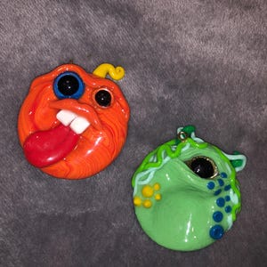 May include: Two circular clay pendants. The left pendant is orange with a yellow stem, large black and blue eyes, white teeth, and a protruding red tongue. The right pendant is light green with a dark eye, blue dots, and green wavy lines.