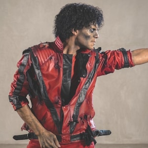Puede incluir: Una chaqueta de piel sintética roja y negra con un diseño de panel frontal en forma de V, cuello alto y varias correas negras con hebillas en la cintura. La chaqueta presenta detalles desgastados, incluyendo pequeños desgarros y bordes deshilachados.