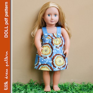 May include: A doll wearing a blue and brown floral print dress with blue straps and buttons. The doll has blonde hair and blue eyes.