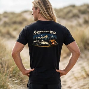 Puede incluir: Camiseta negra con un diseño gráfico que presenta un coche deportivo blanco, una fogata y montañas bajo un cielo estrellado. El texto "Supercar in the Wild" está arqueado sobre el coche. La persona de la imagen lleva la camiseta.