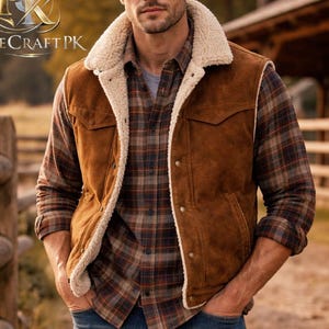 Puede incluir: Un chaleco de ante marrón con cuello y forro de borrego color crema. Se lleva sobre una camisa a cuadros y vaqueros azules. La imagen está ambientada al aire libre, con una valla de madera al fondo.